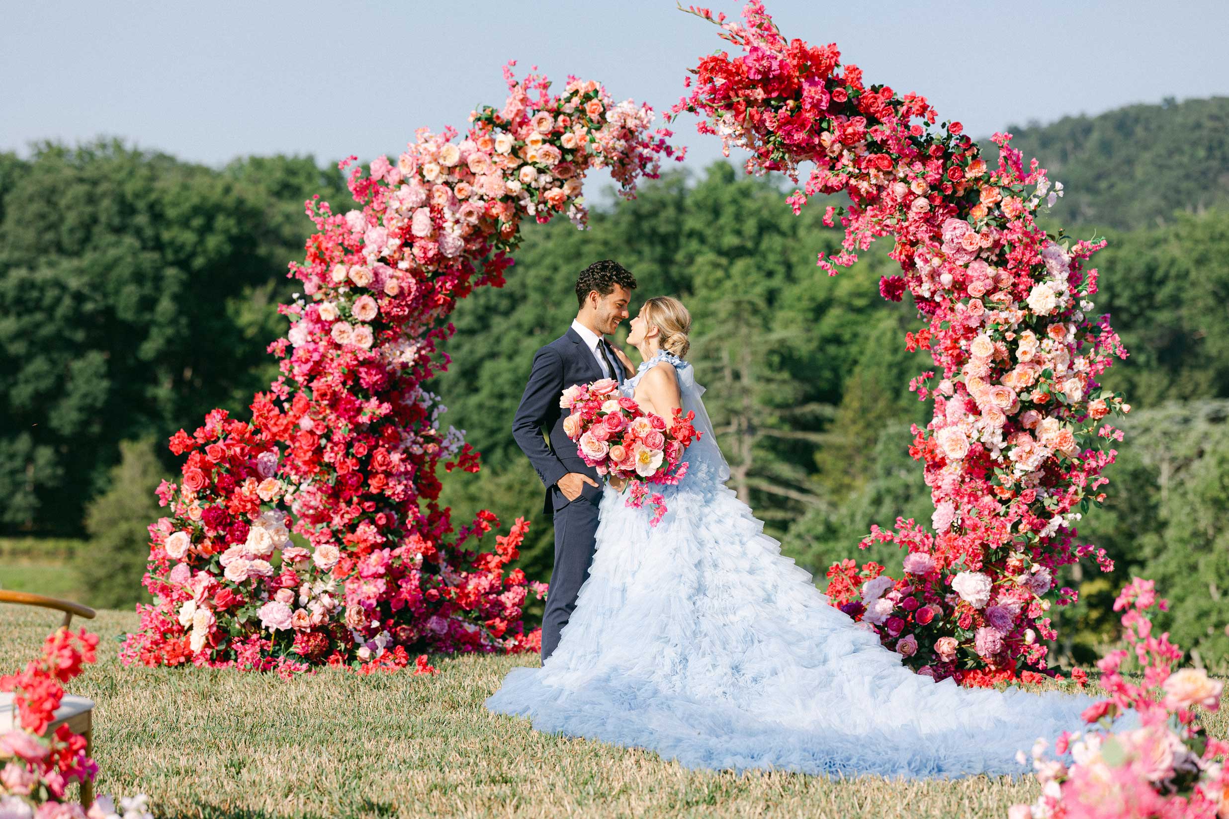 The happy couple poses in front of a stunning floral arch, the bride holding a bouquet and the groom looking dapper in a suit. Timeless Wedding Photography.