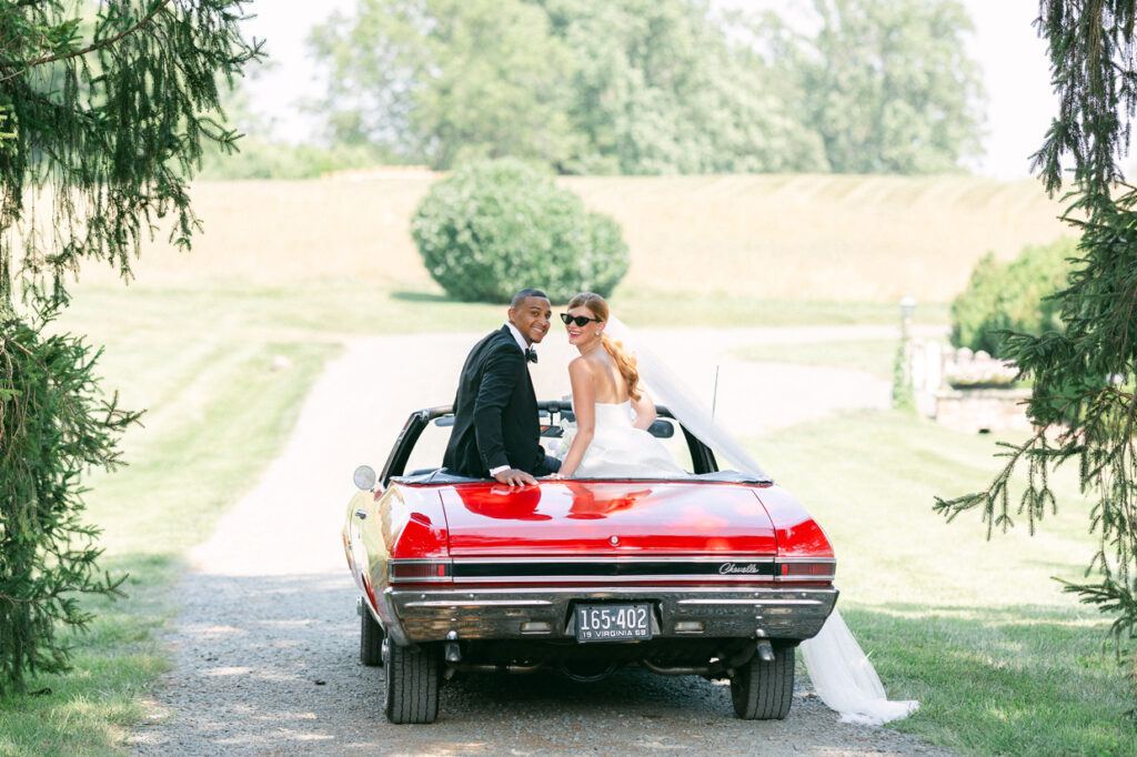 Newlyweds stand in the rear of a crimson vehicle, the bride in a white gown and the groom in a black suit.
