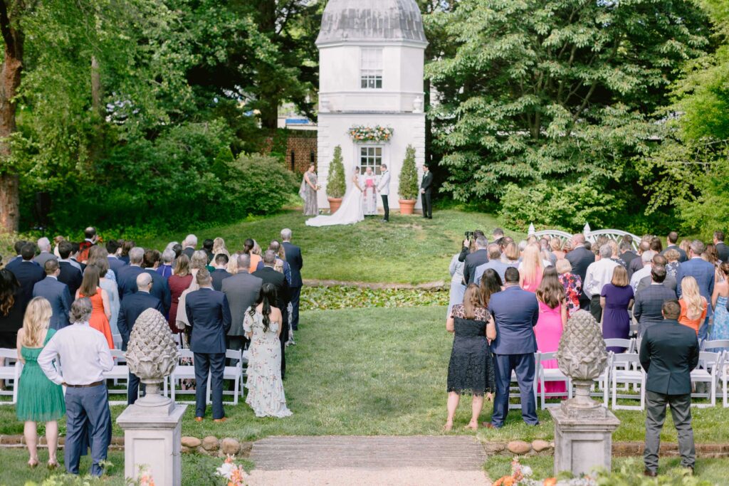 A formal wedding ceremony taking place in a garden with a tower in the background. Guests stand with the couple as they exchange vows.
