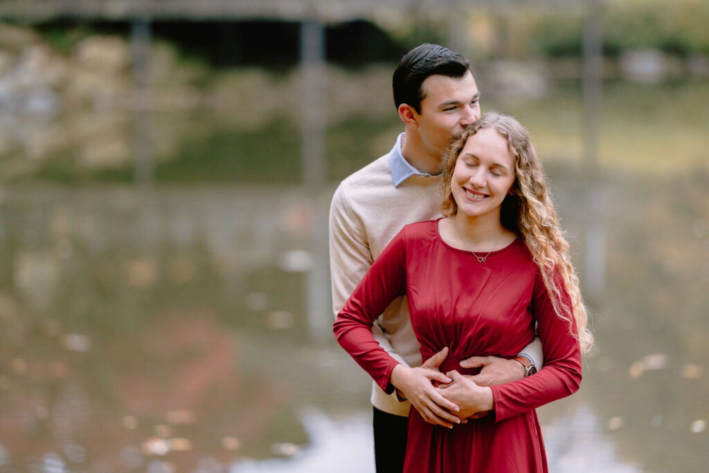 A couple dressed in red stands by a serene lake, celebrating their engagement session in fall at the brookside gardens. The man embraces the woman from