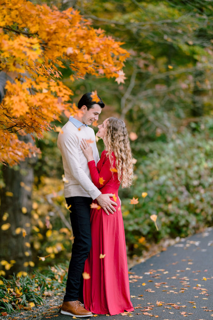 A couple poses amidst stunning autumnal scenery in a romantic fall engagement session, with leaves falling around them. Picture taken by Get ready Photo in the Brookside Gardens. 