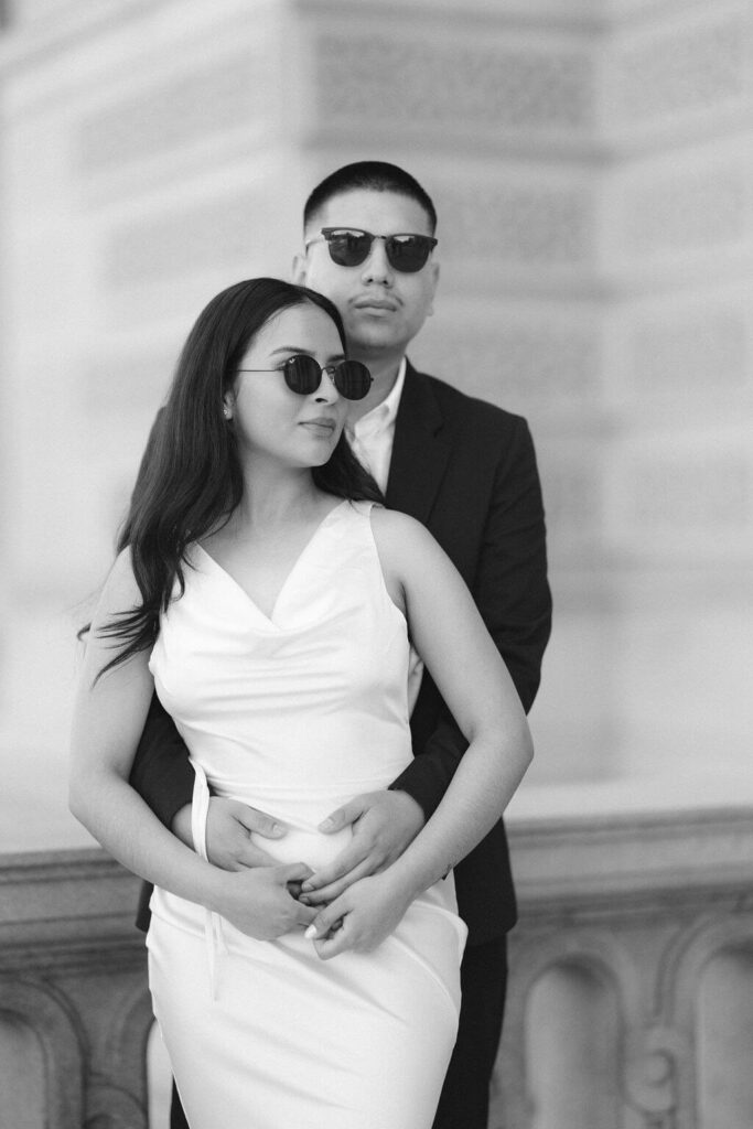 A black and white photo capturing a couple's serene moment on the steps of a grand building in Washington DC, taken by Get Ready Photo. The man stands behind the woman, both dressed elegantly, looking ahead with a shared vision of their future.