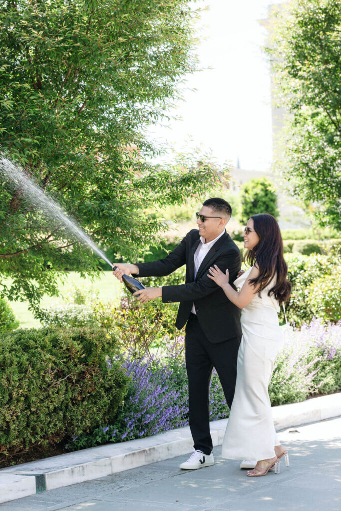 A joyful couple during their engagement session in Washington DC, with the man popping a champagne bottle and the woman laughing beside him. They're stylishly dressed, him in a black suit and her in a white dress, commemorating the moment in a lush garden. This 'Get Ready Photo' captures the excitement and elegance of their special milestone.