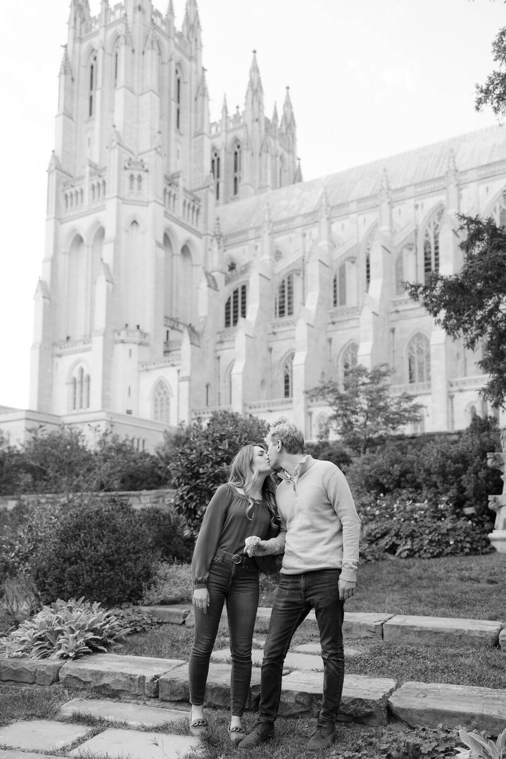 A black and white photo capturing a couple in a sweet embrace, with the iconic Washington National Cathedral towering in the background. The couple stands on a garden path, surrounded by lush greenery, symbolizing the growth and strength of their relationship. This image is part of an engagement session by Get Ready Photo, highlighting the beauty of Washington DC
