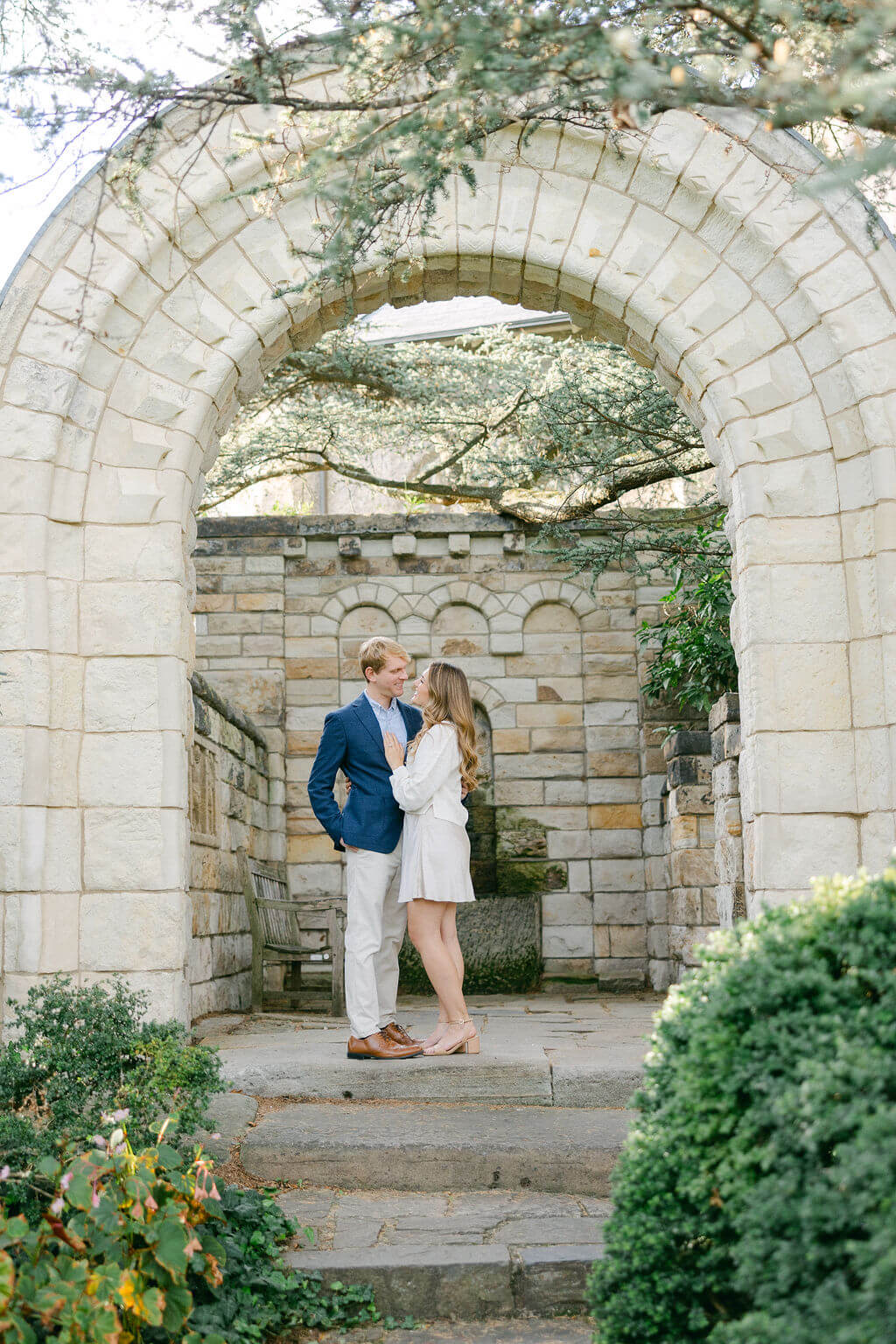 A photo of a couple standing under an arched stone gateway, sharing an intimate moment during their engagement session at the Washington National Cathedral. The historic architecture and serene greenery provide a romantic backdrop, representing their journey ahead. Captured by Get Ready Photo, Washington DC wedding photographers.