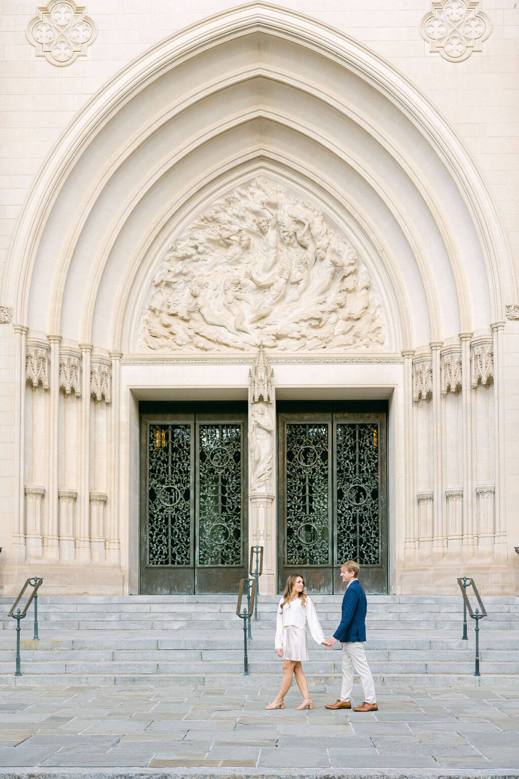 A photo of an engaged couple holding hands in front of the grand entrance of the Washington National Cathedral, with intricate gothic details and a bas-relief above the doors, captured by Get Ready Photo.