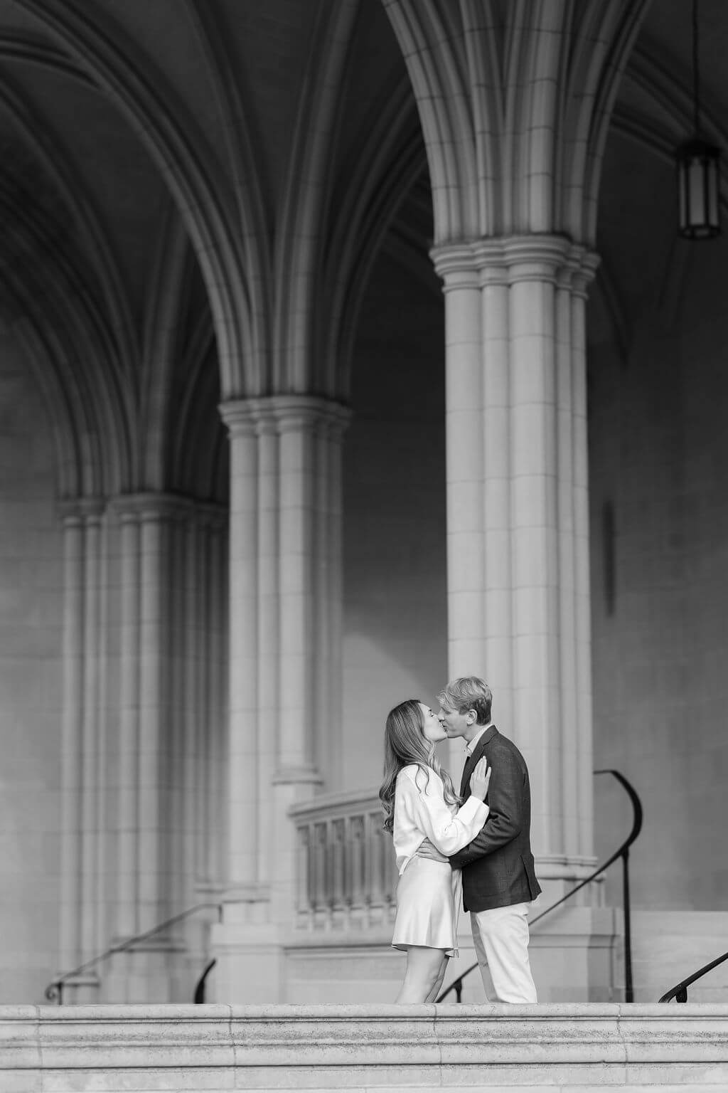A black and white photo of an engaged couple kissing on the steps of the Washington National Cathedral, framed by elegant arches, captured by Get Ready Photo