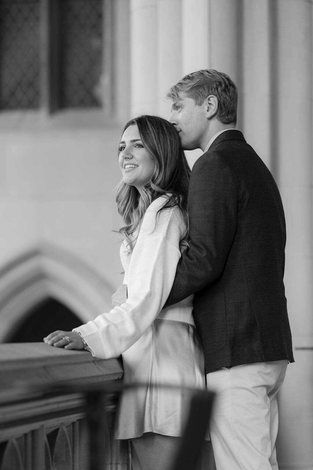 A black and white photo of a smiling woman leaning on a balcony with a man embracing her from behind, both looking into the distance, set against the architectural backdrop of the Washington National Cathedral, captured by Get Ready Photo