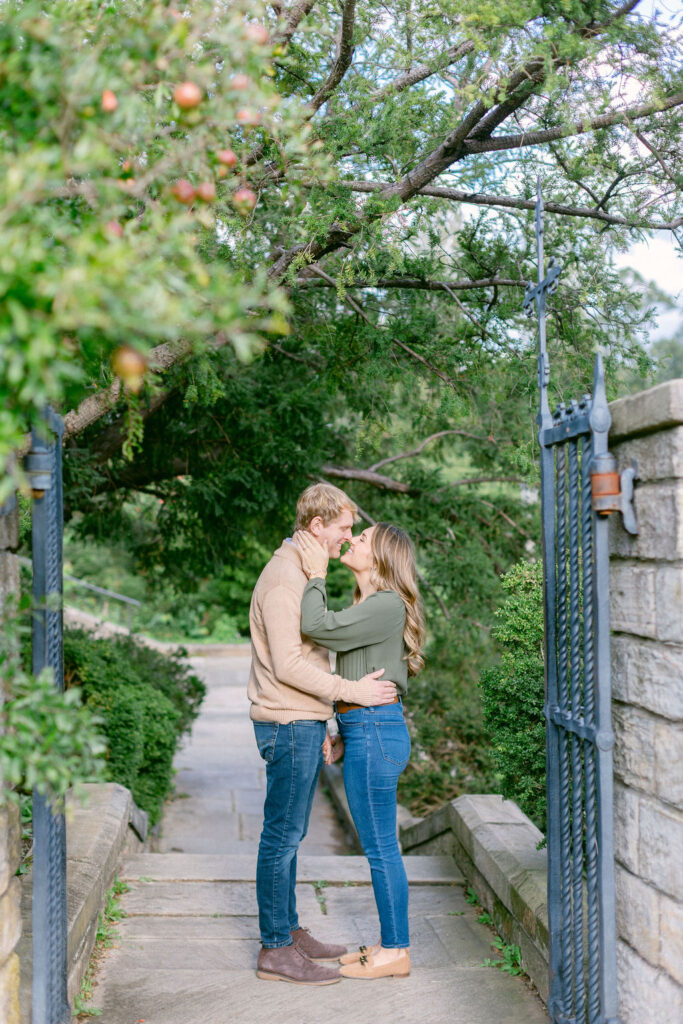 A photo of an engaged couple sharing an intimate moment on a stone pathway at the Washington National Cathedral. The man, wearing a beige sweater and blue jeans, gently holds the woman's face as they are about to kiss. The woman, in a green blouse and blue jeans, smiles while holding the man's hand. Lush greenery surrounds them, highlighting the romance of their engagement session with Get Ready Photo.