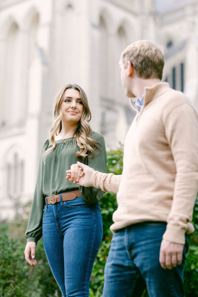 A portrait of a couple embracing, with a natural backdrop of trees at the Washington National Cathedral. The man, in a beige sweater over a blue shirt and jeans, stands behind the woman, who is wearing a green blouse and blue jeans. Both are smiling at the camera, capturing the joy of their engagement session photographed by Get Ready Photo, based in Washington DC