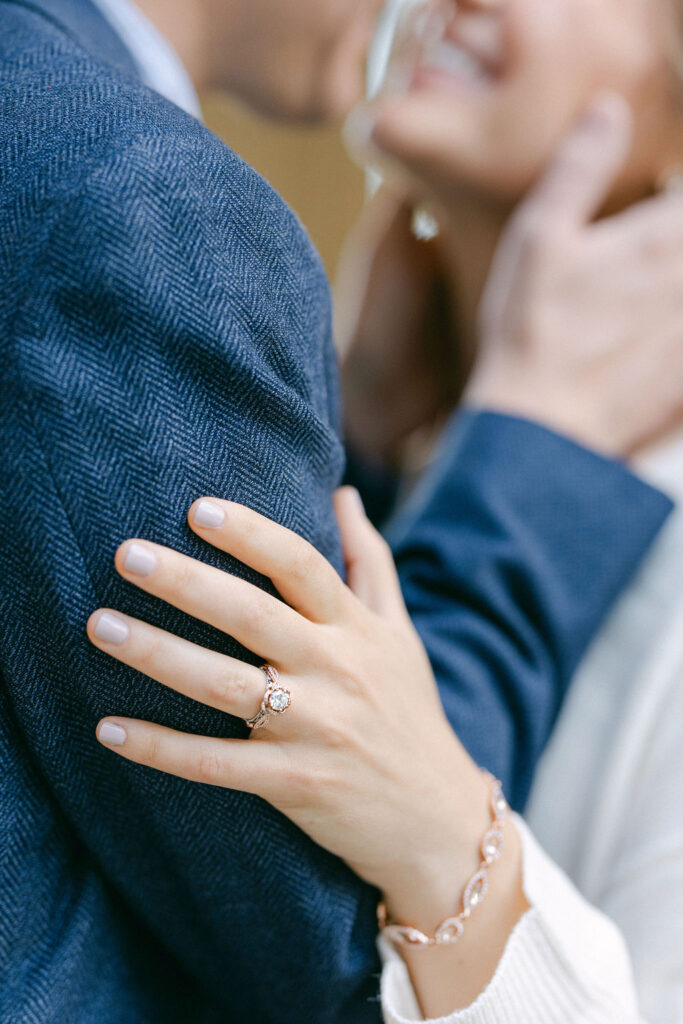 A close-up photo of a woman's hand resting on a man's shoulder, showcasing an engagement ring with a prominent central stone surrounded by smaller diamonds on a gold band. The woman is also wearing a rose gold bracelet with circular, diamond-studded links. Both are wearing blue and white clothing, adding a subtle contrast to the image. The focus on the engagement ring signifies the couple's commitment, suitable for an engagement session by Get Ready Photo, Washington DC wedding photographers.