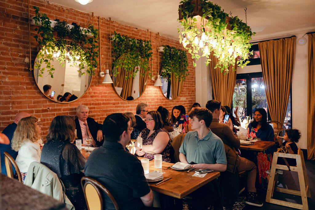 Warm and intimate rehearsal dinner scene in a rustic Washington DC restaurant, photographed by Get Ready Photo, featuring guests engaging in lively conversation surrounded by elegant brick walls and lush greenery.